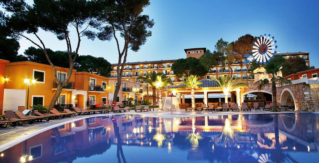 Occidental Playa de Palma hotel in Mallorca, showing its outdoor swimming pool, palm trees and illuminated hotel grounds at dusk, with accommodation reservations available through SIXTY/8 cycling holidays.