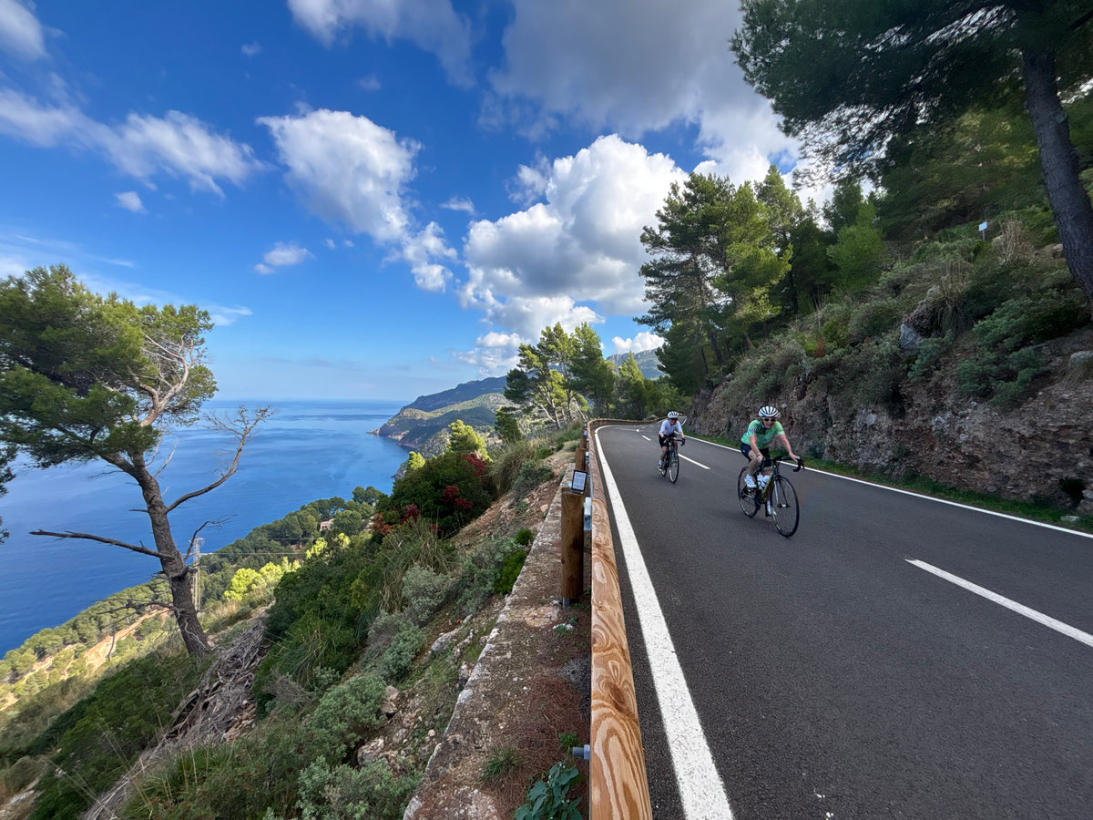 Road cyclists riding along a scenic coastal route in Mallorca, overlooking the Mediterranean Sea, featured in SIXTY/8 cycling holidays and transfer services.