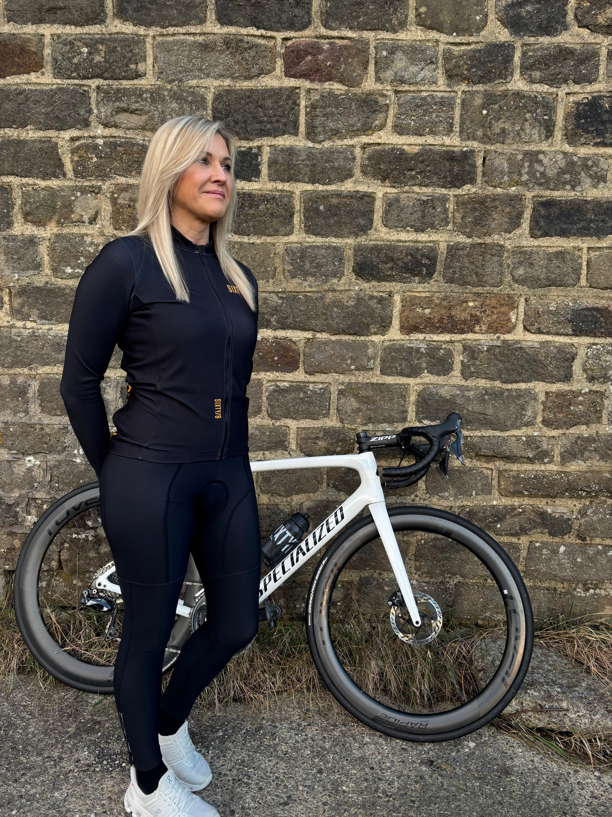 Woman wearing SIXTY/8 black full-length cycling bib tights standing beside a road bike against a stone wall.